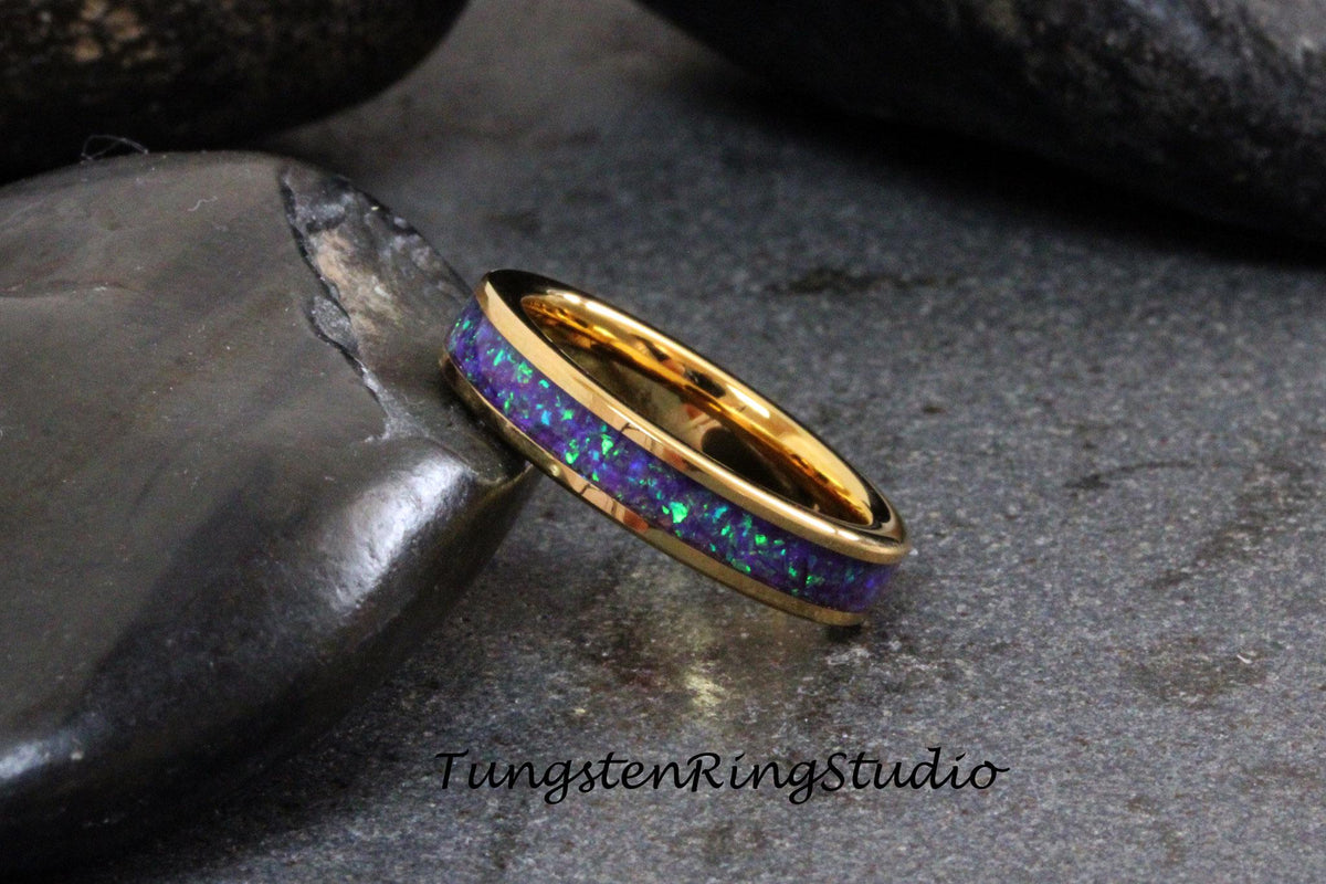 May include: A gold tungsten carbide ring with a purple and green opal inlay. The ring is on a dark gray background.