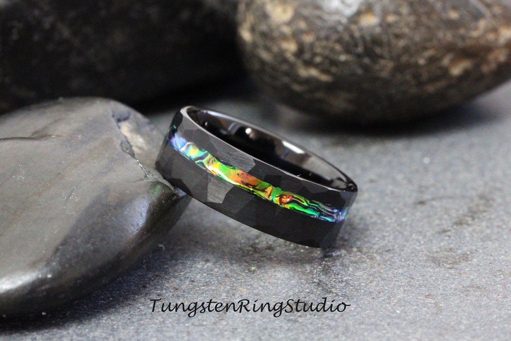 May include: Black tungsten ring with a hammered finish and a thin iridescent abalone inlay. The ring is shown on a dark gray background.