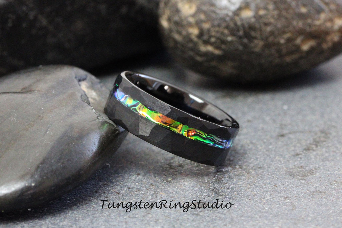 May include: Black tungsten ring with a hammered finish and a thin iridescent abalone inlay. The ring is shown on a dark gray background.