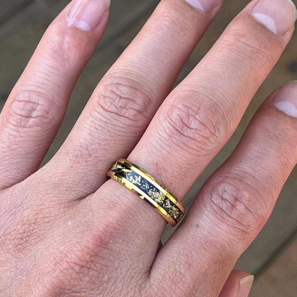May include: A gold-colored ring with a black inlay and gold leaf accents. The ring is shown on a finger, showcasing its design. The band is approximately 0.3 inches wide, featuring a polished finish.