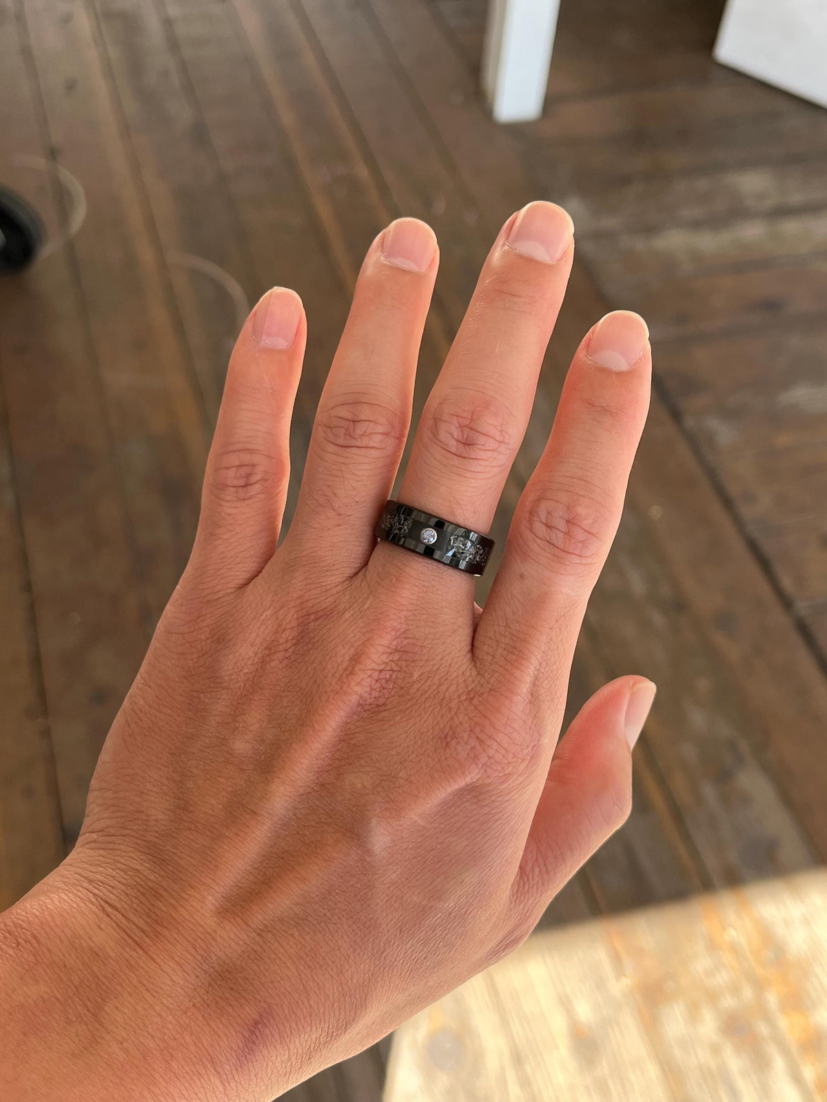 May include: A black band ring featuring a polished finish with a single clear round stone set in the center. The ring has a flat profile and a smooth surface, showcasing a dark metallic appearance that contrasts with the bright, reflective stone.