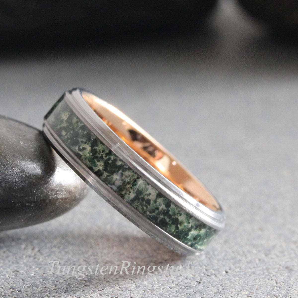May include: A ring featuring a dark grey metallic outer band with a central inlay of mottled green stone and a smooth rose gold interior. The band has a polished finish and a distinct stepped edge design, showcasing a textured green mineral pattern.