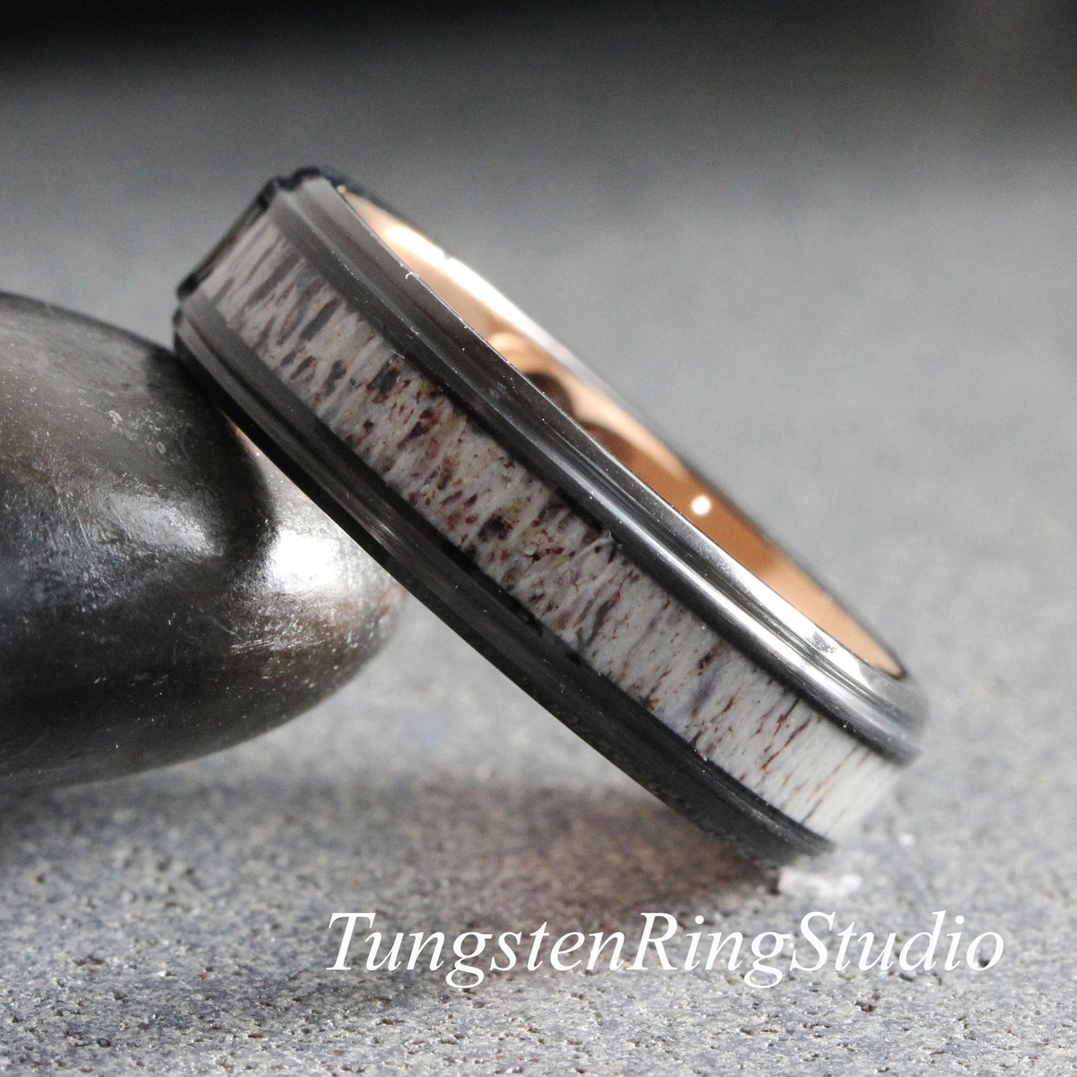 May include: A ring featuring a black metal band with a central inlay of textured, light-colored antler material and a smooth, rose gold-toned interior. The text TungstenRingStudio is visible at the bottom of the image.