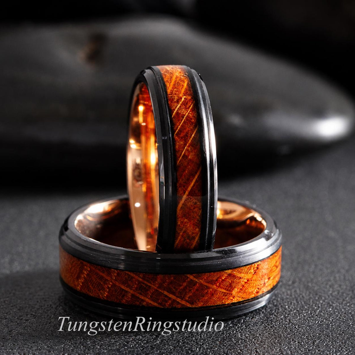 May include: Two rings featuring a black metal band with a central inlay of brown wood grain. The interior of the rings is a polished rose gold tone. The text TungstenRingstudio is visible at the bottom of the image in a white font.